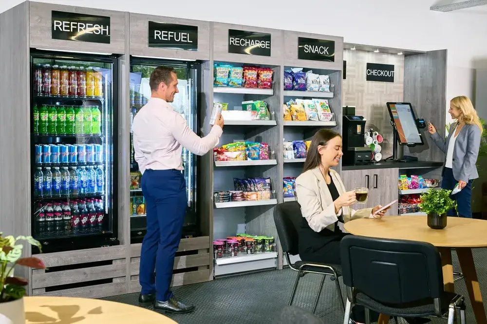 A modern micro-market area in an office setting. A man in a pink shirt is selecting a drink from a cooler labeled REFUEL. Shelves stocked with snacks are labeled "RECHARGE" and "SNACK. A woman sits at a table with a laptop and coffee, while another woman stands at a self-checkout kiosk labeled CHECKOUT.